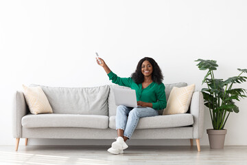 Obraz premium Happy black woman sitting on couch with laptop, holding remote