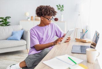 Smart black teenager using tablet and laptop for online studies at home. Domestic schooling concept