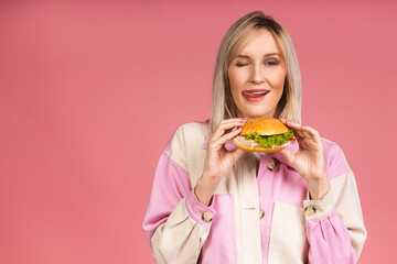 Cheerful young hungry beautiful blonde woman holding cheeseburger or burger holding in hands isolated over pink background. Junk food concept.