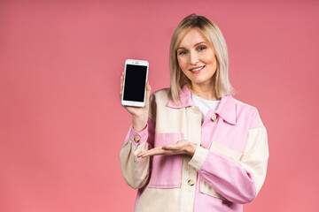 Naklejka premium Portrait of attractive young blonde woman with white smile, showing empty smartphone screen, demonstrate an app or online shopping store, isolated over pink background.