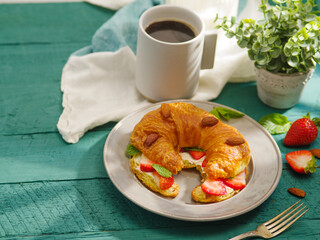 A cup of black coffee, croissant with fresh strawberries, butter, almond nuts, fork, indoor flower. Turquoise background, white napkin. Beautiful composition, menu design, culinary blog.