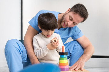 Young therapist helping cute little boy who has cerebral palsy, playing with developing toy at...