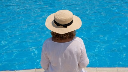 Senior woman relaxing near the hotel's blue outdoor swimming pool. People are enjoying their summer vacation. All inclusive, back view