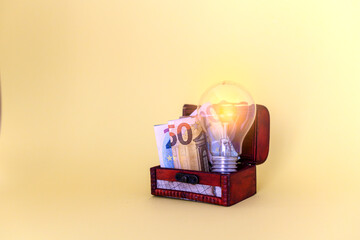 Lit light bulb next to money in euro banknotes inside a trunk. Concept of the high price and rise in the electricity bill.