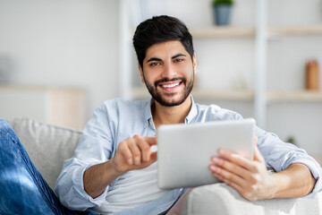 Portrait of excited arab man using tablet, sitting on couch at home and smiling to camera, free space