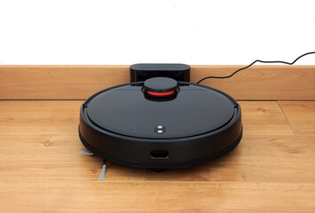 Black robot vacuum cleaner is charging on charging stand. Laminate flooring in modern house....