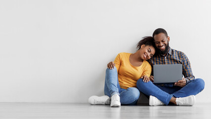 Cheerful young black man and woman look in laptop, planning interior, sits on floor in empty room