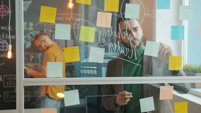 Portrait of young businessman working with glass board writing on sticky notes while male colleague standing near chalkboard and thinking