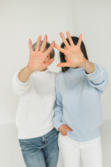 Two women cover their faces with their hands, fingers wide open. White and blue clothes. light background. Brunette and blonde. Long and short hair.
