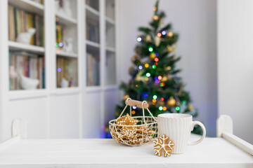 White cup with tea and gingerbread cookies in the form of a snowflake on the table against the background of an unsharp Christmas tree with lights in the interior