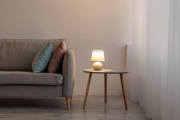 Sofa with pillows, glowing lamp on table on gray wall background in evening in office or living room
