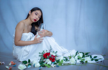 Bella chica asiática sentada con toga sujeta jarra con agua. Suelo cubierto de flores. Fondo azul 