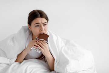 Sad lonely hungry european young lady eating chocolate, wrapped in blanket in bedroom interior