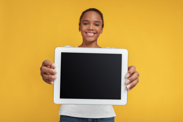Smiling teen african american girl show tablet with empty screen, isolated on yellow background