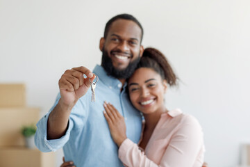 Cheerful young african american husband hugs wife and shows keys in room with cardboard boxes