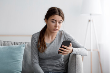 Sad young female sits on sofa look at smartphone and reading message at home, frustrated by received notification