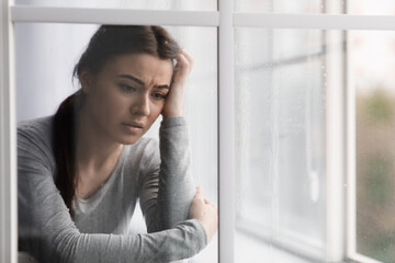 Unhappy sad european young lady suffering from depression, stress and mental problems near window with rain drops
