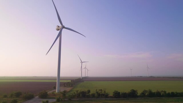 Windmills in a green field at sunset. Wind farm with turbine cables for wind energy. Renewable energy source, earth care. Drone video of wind power station, 4k. Botiyevska Ves, Ukraine.