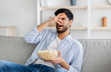 Young arab man watching scary movie, closing eyes in horror, eating popcorn, resting on sofa at home, free space
