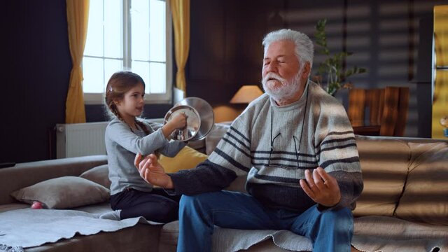 Senior man meditating sitting on sofa while active child granddaughter playing with lids. Child bothering mindful grandfather do yoga exercise relax lotus pose