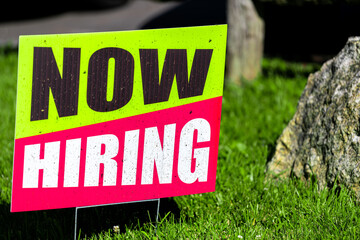 Sign text closeup for help wanted now hiring in English with red and green colors as signpost on grass for store shop business during corona virus covid 19 pandemic