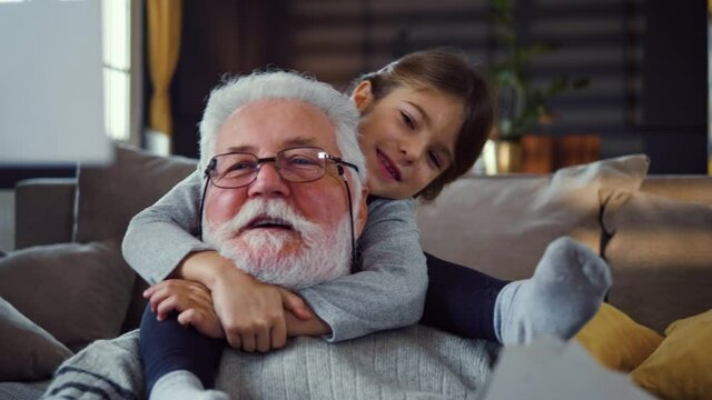 Happy moments of a child girl and a grandfather at home while looking at pictures together.  Granddaughter hugs the old man looking together at his pictures from his youth. 