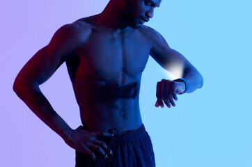Cropped view of handsome young black guy with naked torso checking fitness tracker in neon light