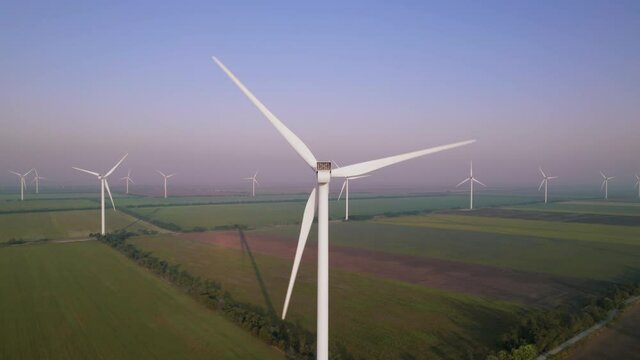 Windmills with rotating wings among green fields. Wind farm with turbine cables for wind energy. Renewable energy source, earth care. Drone video of wind power station, 4k. Botiyevska Ves, Ukraine.