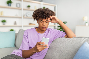 Funky black teen guy with mobile phone studying online, messaging with friend, posting in social network from home