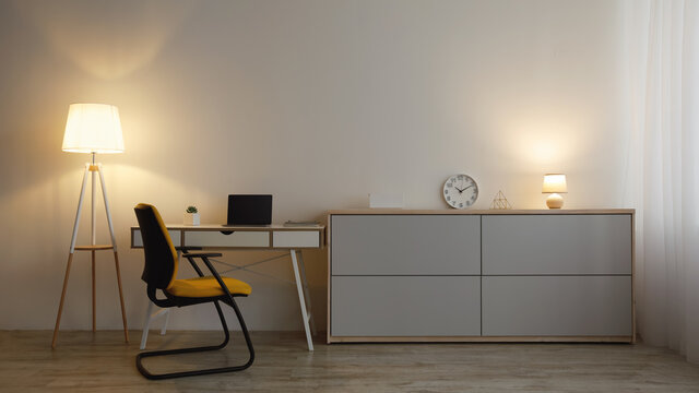 Comfortable armchair, table with laptop with empty screen, furniture with clock, glowing lamps on gray wall background