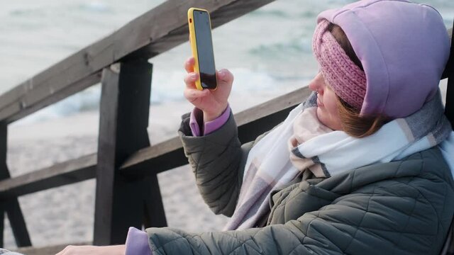 Woman blogger in warm clothes does selphie on the winter beach. Traveller at the cold sea in the off-season, 4k 60p