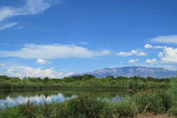 Distant Mountain Landscape against Bright Blue Sky with Fluffy White Clouds and Peaceful Lake in Foreground