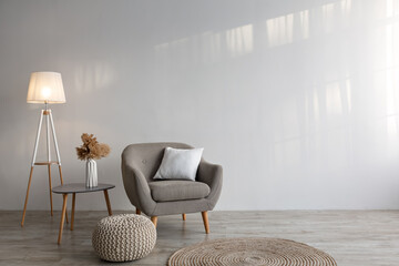 Cozy armchair with pillow, luminous lamp, table, dry plants in vase, ottoman and carpet on floor on gray wall background