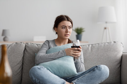 Upset apathetic european millennial woman sits on sofa with glass and suffering from depression and stress