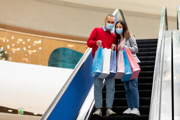 Full length of young multiracial couple in face masks going down on moving stairway at mall, looking into shopping bags