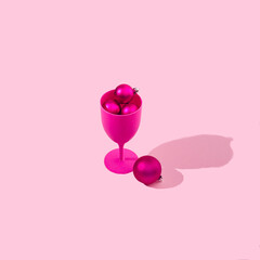 Fototapeta premium A fuchsia wine or cocktail glass full of fuchsia Christmas baubles on pastel pink background. Monochrome colors for Christmas or New Year celebration banner or advertisement.