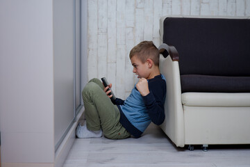 mały chłopak siedzi między fotelem a szafą, trzyma w dłoni telefon jest zdenerwowany - obrazy, fototapety, plakaty