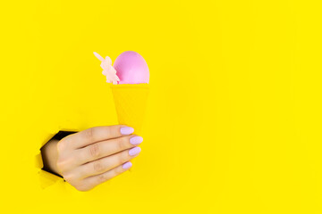 Abstract yellow background with female hand is holding pink egg in colorful ice cream cone. Minimal...