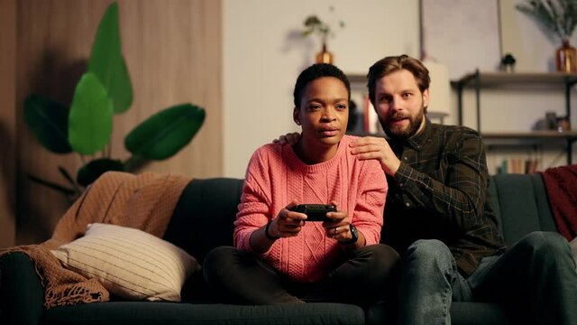 Portrait of young beautiful gamer African American girl sitting on sofa and playing in video games on digital console. A guy who supports and encourages his girlfriend. High quality 4k footage