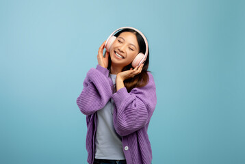 Excited asian woman listening to music with closed eyes using wireless headphones, standing over...