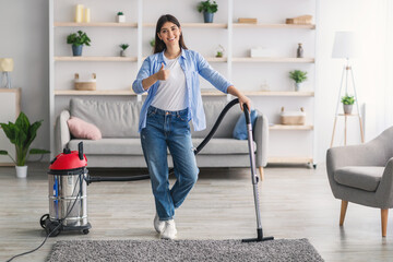 Fototapeta na wymiar Happy woman cleaning carpet with vacuum cleaner, showing thumbs-up