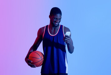 Cheerful young black basketball player holding ball and using smartphone in neon light
