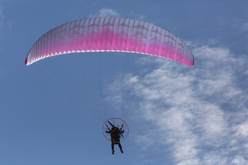A man flies a motorized pink and white parasail away from the camera through a blue sky with scattered puffy clouds.