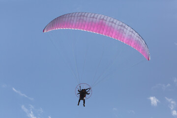 A man flying a pink and white motorized parasail moves away from the camera through a blue sky with a few bits of puffy white cloud above him.