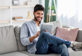 Portrait of happy arab guy using digital tablet, resting on couch in living room interior, free...