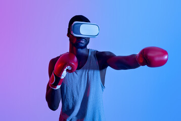 Young black guy in VR headset wearing boxing gloves, making punch in neon light. Exercising with...