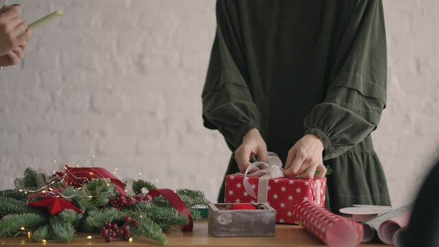 Crop hands decorating wrapped box with string. Female packing cardboard gift box on wooden table with various decorative items prepared for Christmas celebration. Detail female hands tying 