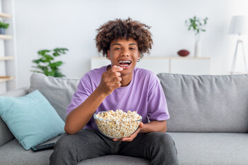 Cheery African American teenager with bowl of popcorn enjoying funny movie, laughing at home