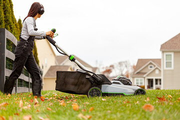 a young caucasian woman wearing overalls and noise blockers is operating a lawnmover on grass in late autumn. There are fallen leaves and a suburban neighborhood setting in background.