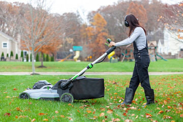 a young caucasian woman wearing overalls and noise blockers is operating a lawnmover on grass in late autumn. There are fallen leaves and a suburban neighborhood setting in background. - obrazy, fototapety, plakaty
